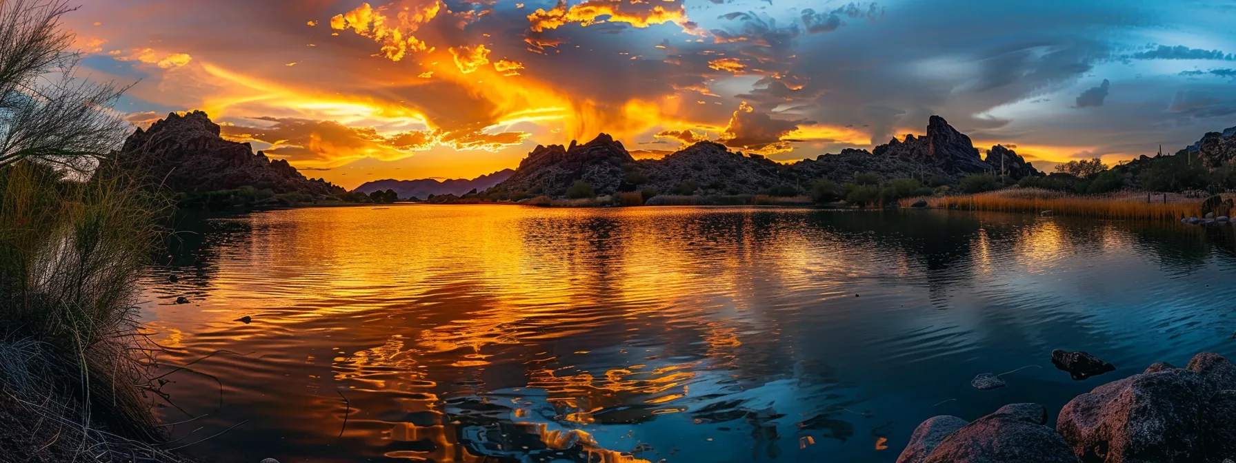 a vibrant sunset casts a warm glow over a tranquil lake, with silhouetted mountains framing the horizon and fluffy clouds reflecting golden hues on the water's surface.